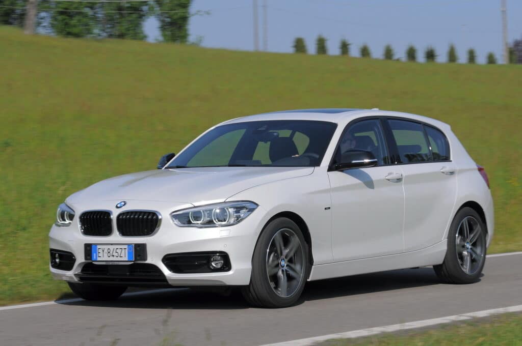 Los 12 mejores coches de segunda mano por menos de 10.000 euros 5 BMW Serie 1 116d F20 de color gris conducido por un hombre en una carretera con paisaje verde, pinos de fondo.