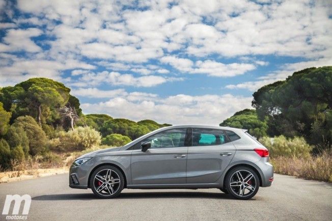 Los 12 mejores coches de segunda mano por menos de 10.000 euros 11 SEAT Ibiza 1.0 TSI de color gris cruzando en medio de la carretera, paisaje de árboles verdes y cielo seminublado.