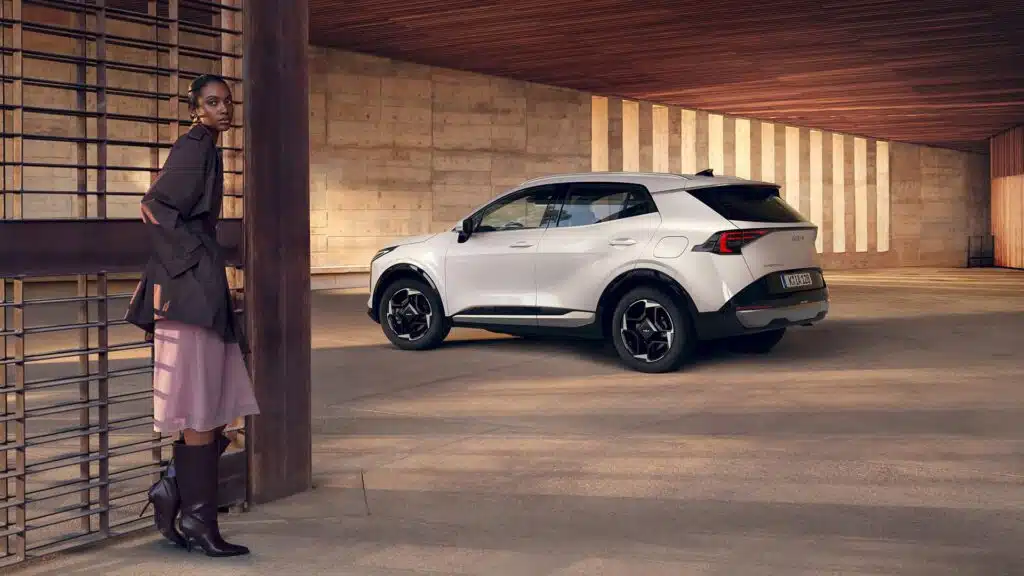 Los 8 SUV más fiables en 2026. Cuál deberías comprar 5 Mujer con saco marrón, falda rosa y botas marrones hasta las rodillas apoyada sobre una reja, de fondo se ve un Kia Sportage de color blanco.
