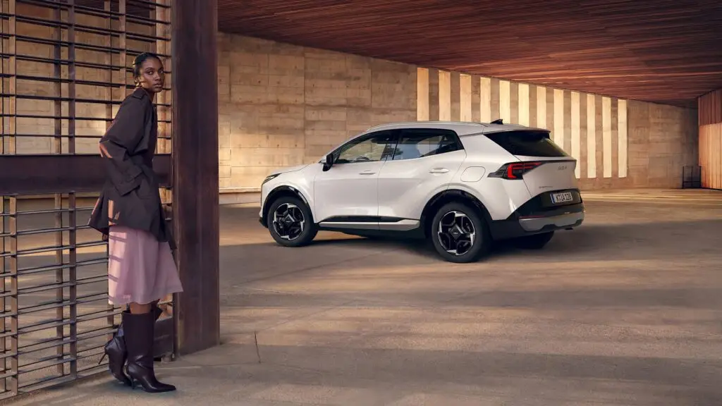 Los 8 SUV más fiables en 2026. Cuál deberías comprar 5 Mujer con saco marrón, falda rosa y botas marrones hasta las rodillas apoyada sobre una reja, de fondo se ve un Kia Sportage de color blanco.