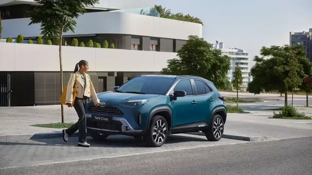 Los 8 SUV más fiables en 2026. Cuál deberías comprar 8 Mujer pasando por delante de un Toyota Yaris Cross celeste estacionado en una zona urbana.