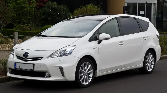 Los 24 mejores coches familiares de 7 plazas de ocasión 8 Toyota Prius+ blanco con el distintivo Hybrid en la aleta, aparcado de tres cuartos delantero ante un edificio