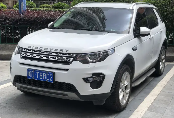 Los 24 mejores coches familiares de 7 plazas de ocasión 24 Land Rover Discovery Sport blanco aparcado sobre una acera, de tres cuartos delantero con un seto verde detrás